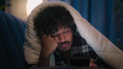 Sad sleepy guy Latino tired man upset Arabian sleeping napping boring Indian male at night evening dark home under blanket cover with duvet scrolling mobile phone addict smartphone bored social media