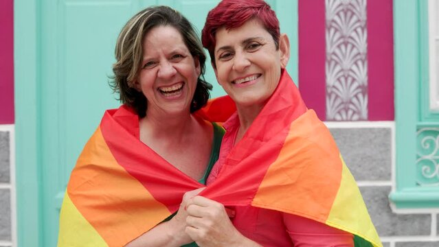 Happy Senior gay lesbian couple having fun wearing Lgbtq rainbow flag at pride parade - Family and love concept - Focus on right woman face