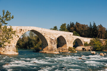 Flowing river beneath the historic Aspendos bridge, surrounded by lush vegetation and the timeless...