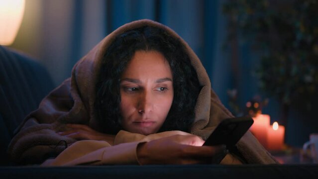 Hispanic sad sleepy girl Latino tired woman upset boring Indian lady bored Arabian female night evening dark home under blanket cover with duvet scrolling mobile phone addict smartphone social media