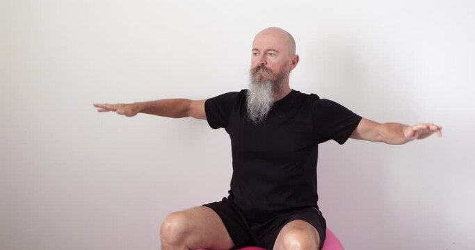 50 year old mid adult man with long beard sitting on fitness ball, doing neck and upper spine exercise at home, lifting arms upwards and downwards, real people