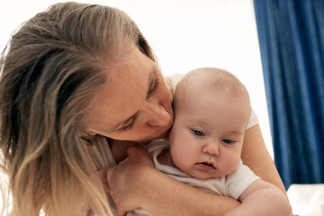 Mom hugs and kisses her little child in a bright child's room. Mom carefully holds the baby in her arms. Happy childhood