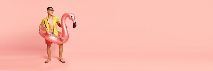 Banner with handsome young student, man wearing beach clothes standing with inflatable flamingo over pink background. Copy space for ad - obrazy, fototapety, plakaty
