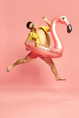 Portrait with attractive young guy, man wearing beach clothes jumping with inflatable flamingo over...