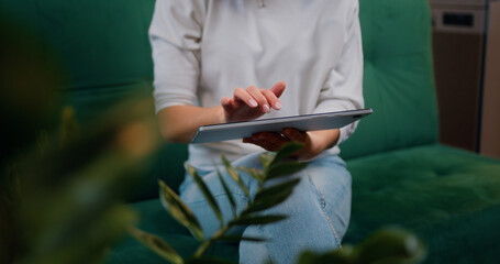 Close-up woman holding digital tablet in hands and using business apps typing surfing the web or using social networking sites while sitting on green sofa at home