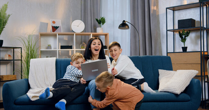 Good-looking happy smiling mother sitting on the couch with laptop when three playful different ages sons running up to her with huggs,family concept
