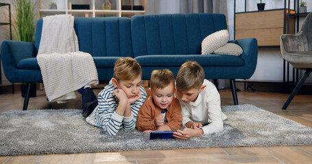 Lovely smiling happy different ages three boys lying on the floor and using smartphone to watch...