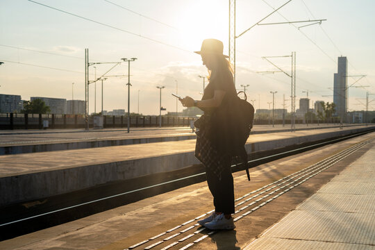 Tourist woman with backpack standing on railway station platform and using phone at sunset.