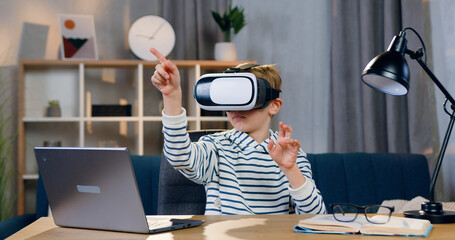 Portrait of attractive smart serious 10-aged boy in virtual reality headset which sitting at his workplace and working on imaginary screen