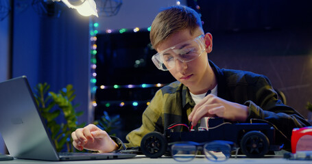 Smart teenage schoolboy using laptop and carefully disassembling his remote controlled toy car homework project at home. People with futeristic technology and education concept.