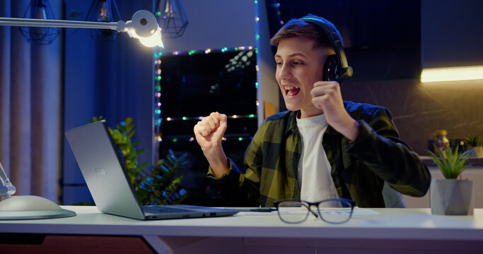 Teenage boy sits at desk using laptop reading e-mails online, gets good news, positive exam results and celebrates victory in evening at home.