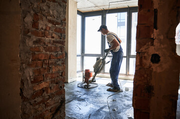 Male worker using troweling machine while screeding floor in apartment under renovation. Man...