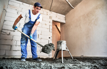 Man preparing floor screed material while standing near concrete screed mixer machine. Male worker using shovel while shoveling sand-cement mix in building under construction.