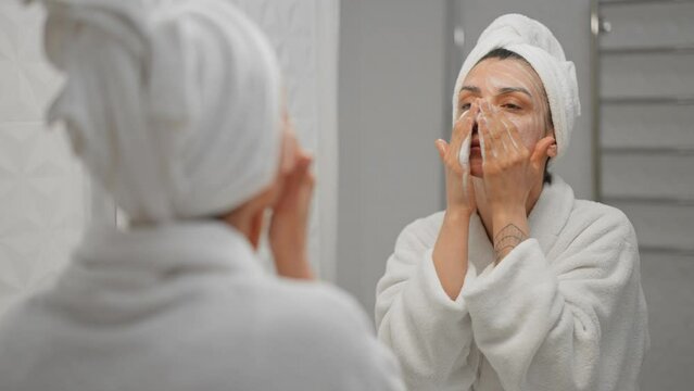 Woman using moisturizing foam for face washing and smiling, morning skincare routine. Young female student wearing towel on head looking in mirror in spa salon. Preparation for make up