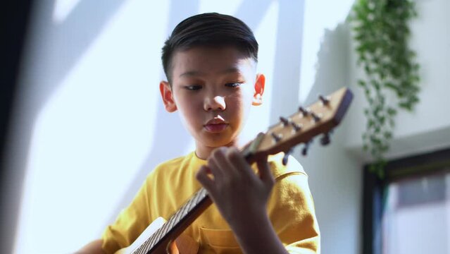 Preteen Asian boy playing acoustic guitar dressed sitting on the cozy sofa at the home living room and enjoying a favorite hobby, Music education concept image.