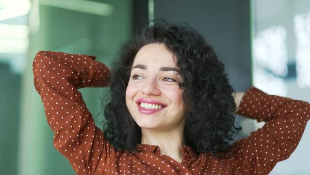 Close up portrait of a pretty smiling businesswoman resting while sitting in a modern office looking away. A curly cheerful brunette sits with her hands behind her head. Positive happy female employee
