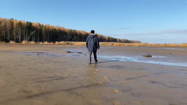 a man in a hat and jacket walks along a damp sandy beach in autumn.