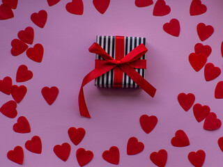 Wrapped gift box is on red heart paper background, top view. Greeting card, present. Valentines day holiday concept. Flat lay with a lot of tiny heart-shaped papers with space for text. February 14th.
