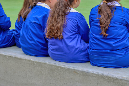 Three Female Students In Blue Aprons From The Italian Elementary School Seen From Behind, Sitting On A Low Wall