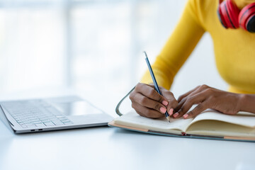 Close-up of a woman taking notes on notebook with laptop in office.