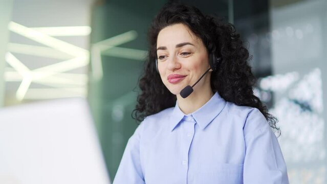 A brunette woman in a headset customer service operator talking on a video call while sitting in the office. Call center agent advises the client by answering complaints and questions using a laptop