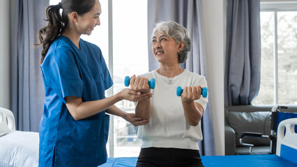 Young physiotherapist helping senior mature asian woman grey hair work out with dumbbells, to...