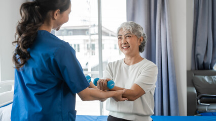 Fototapeta premium Young physiotherapist helping senior mature asian woman grey hair work out with dumbbells, to recover from injury at health centre in physical therapy session.