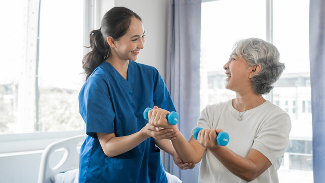 Young physiotherapist helping senior mature asian woman grey hair work out with dumbbells, to recover from injury at health centre in physical therapy session.