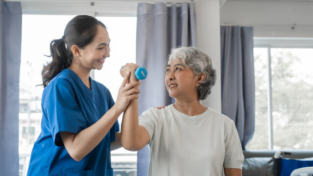 Young physiotherapist helping senior mature asian woman grey hair work out with dumbbells, to recover from injury at health centre in physical therapy session.