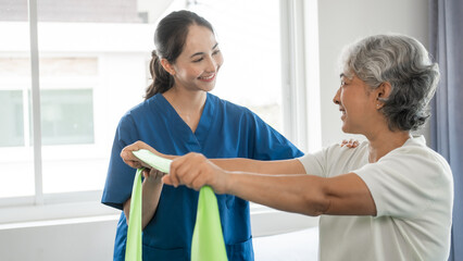 Obraz premium Young physical therapist caregiver assisting mature asian woman grey hair doing exercise with elastic bands at physiotherapy clinic.