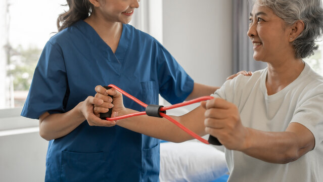 Young physical therapist caregiver assisting mature asian woman grey hair doing exercise with elastic bands at physiotherapy clinic.