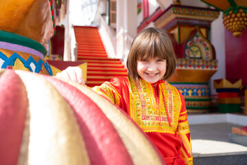 a happy boy in a Russian national bright red-orange costume near the Russian national multi-colored Terem building	