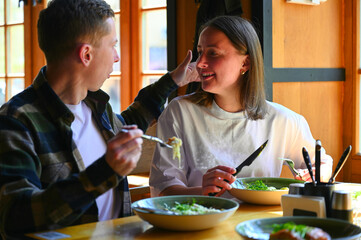 Sitting by a bright window in a cozy restaurant, a couple shares a meal together during lunchtime. The natural light illuminates the space, they chat and enjoy their food.