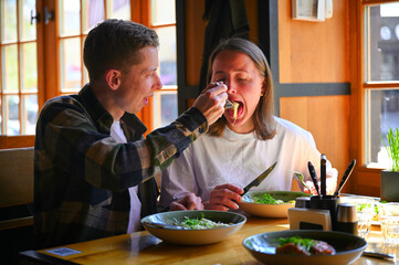 Sitting by a bright window in a cozy restaurant, a couple shares a meal together during lunchtime. The natural light illuminates the space, they chat and enjoy their food.