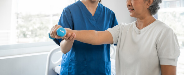 Young physiotherapist helping senior mature asian woman grey hair work out with dumbbells, to recover from injury at health centre in physical therapy session.