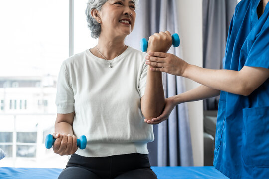 Young physiotherapist helping senior mature asian woman grey hair work out with dumbbells, to recover from injury at health centre in physical therapy session.