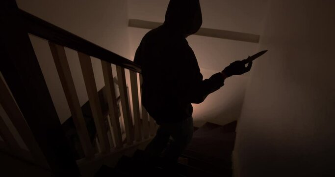 A thief with a pocket knife sneaks into the house quietly down the stairs at night.