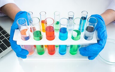 Fototapeta premium Row of colorful liquid chemical in test tube with scientist hand holding while wearing rubber blue gloves with laptop keyboard and sample tray in white lab
