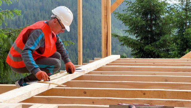 Carpenter in the process of building wooden frame house. Man measures distances with a measuring tape while dressed in workwear and helmet. Concept of modern, environmentally-friendly construction.