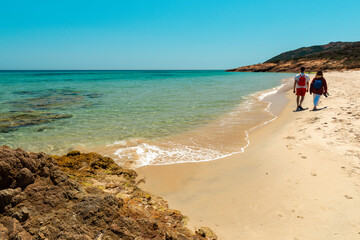 Couple on the beach walking in a peaceful Mediterranean holiday location with calm and turquoise sea. Spiaggia di Santa Margherita di Pula, Sardinia, Italy
