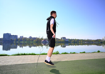 Man jumps rope near the lake in the morning, dressed in sportswear and looking focused on his fitness routine. 