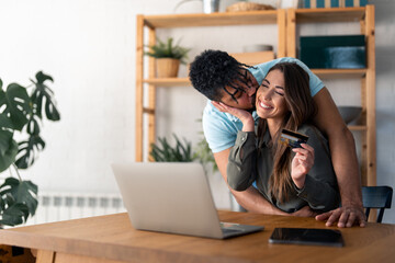 Smiling wife holding credit card while husband is kissing her. Young married couple making online reservations using credit card and laptop technology.