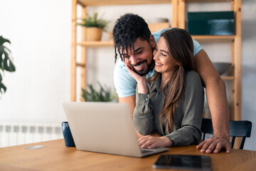 Young smiling couple in love working together on laptop computer. Supportive husband helping his wife with online work. Happy young woman caressing her loving husband while using laptop at home.