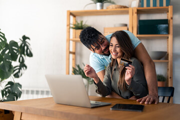 Husband hugging his wife while she's using laptop and credit card at home.