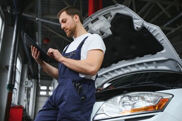 Bearded mechanic in overalls standing in garage of a car salon and holding tablet. He is about to...