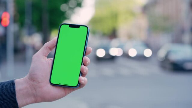 Man's Hand Holding Phone With Green Mock-up Screen On Background of Car Traffic