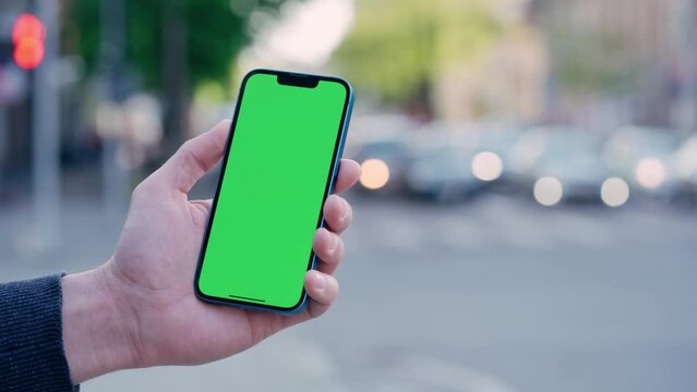 Man's Hand Holding Phone With Green Mock-up Screen On Background of Car Traffic