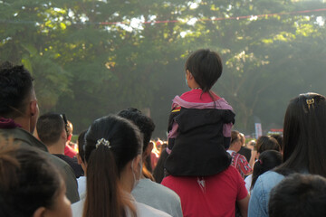 Malaybalay, Philippines - father carries his son, a young boy with a rattail, on his shoulders among a busy crowd at the Kaamulan Festival in Bukidnon. Post-pandemic outdoor event. People from behind.