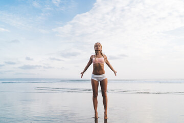 Calm fit girl with closed eyes feeling enlightenment freedom while spending summertime for yoga stretching at seaside on sandy beach, attractive Caucasian woman enjoying healthy lifestyle and healing