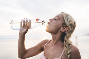 Caucasian female fit girl drinking water for refreshing during workout training at coastline seaside, determined woman with blonde hair holding bottle getting ready for cardio exercising outdoors